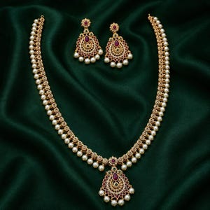 May include: A gold-toned necklace and matching pair of earrings featuring intricate metalwork, teardrop-shaped red stones, small green accents, and dangling white pearls. The jewelry set displays a symmetrical design with ornate floral patterns.