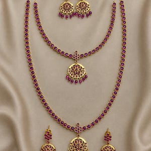 Puede incluir: Un conjunto de joyería compuesto por dos collares dorados y dos pares de pendientes a juego. Cada pieza presenta engastes circulares dorados con incrustaciones de piedras color magenta y pequeñas cuentas colgantes del mismo tono.