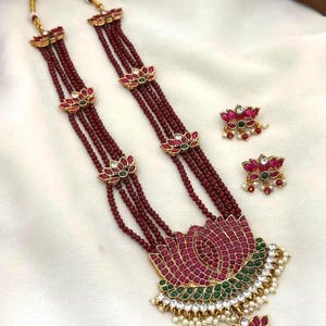 May include: A multi-strand necklace featuring deep red beads and gold-tone lotus flower accents with pink and green stones. The central pendant is a large lotus shape with pink, green, and white stones, accompanied by matching lotus-shaped stud earrings.