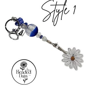 May include: A beaded keychain with a daisy charm, blue and clear beads, and a silver-tone cat charm. The keychain has a silver-tone clasp and a round logo that says "The Beaded Daisy."
