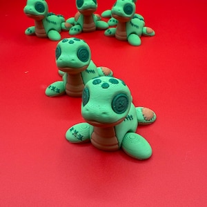 Peut inclure: Plusieurs figurines de tortues vertes et marron, avec des yeux en bouton et des détails cousus, sont disposées sur une surface rouge. Les tortues ont une apparence fantaisiste et artisanale, avec des coutures visibles et des surfaces texturées. L'esthétique générale est ludique et artistique.