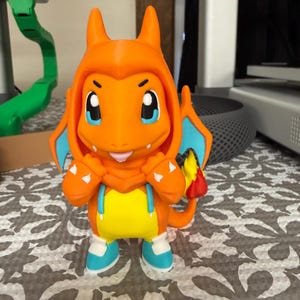 May include: A 3D-printed orange figure of a cartoon character with a hooded outfit. The figure has blue eyes, blue and yellow accents, and a small flame on its tail. The figure is standing on a patterned surface.