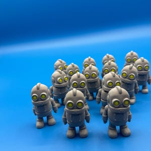 Puede incluir: Un grupo de figuras de robots grises con ojos amarillos se disponen sobre un fondo azul brillante. Los robots tienen un diseño retro, con cuerpos redondeados y detalles sencillos. El tema general es la ciencia ficción o los juguetes.