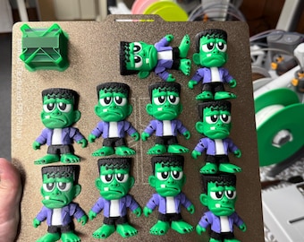May include: A collection of green Frankenstein monster figurines with black hair, black shoes, and purple jackets. The figurines are arranged on a brown surface, with one lying down. A green 3D-printed object is in the upper left corner.