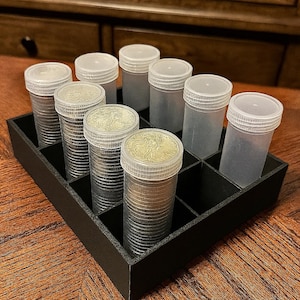 May include: A black, divided tray holding clear plastic tubes filled with silver coins. The tubes have white lids. The tray is on a wooden surface, with a dark wooden cabinet in the background.