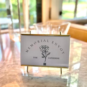 May include: A white sign with gold trim and legs, featuring the text "MEMORIAL ENVOY" in a curved font, and a rose illustration. The names "TOM" and "CATHERINE" are below the rose. A QR code is at the bottom.