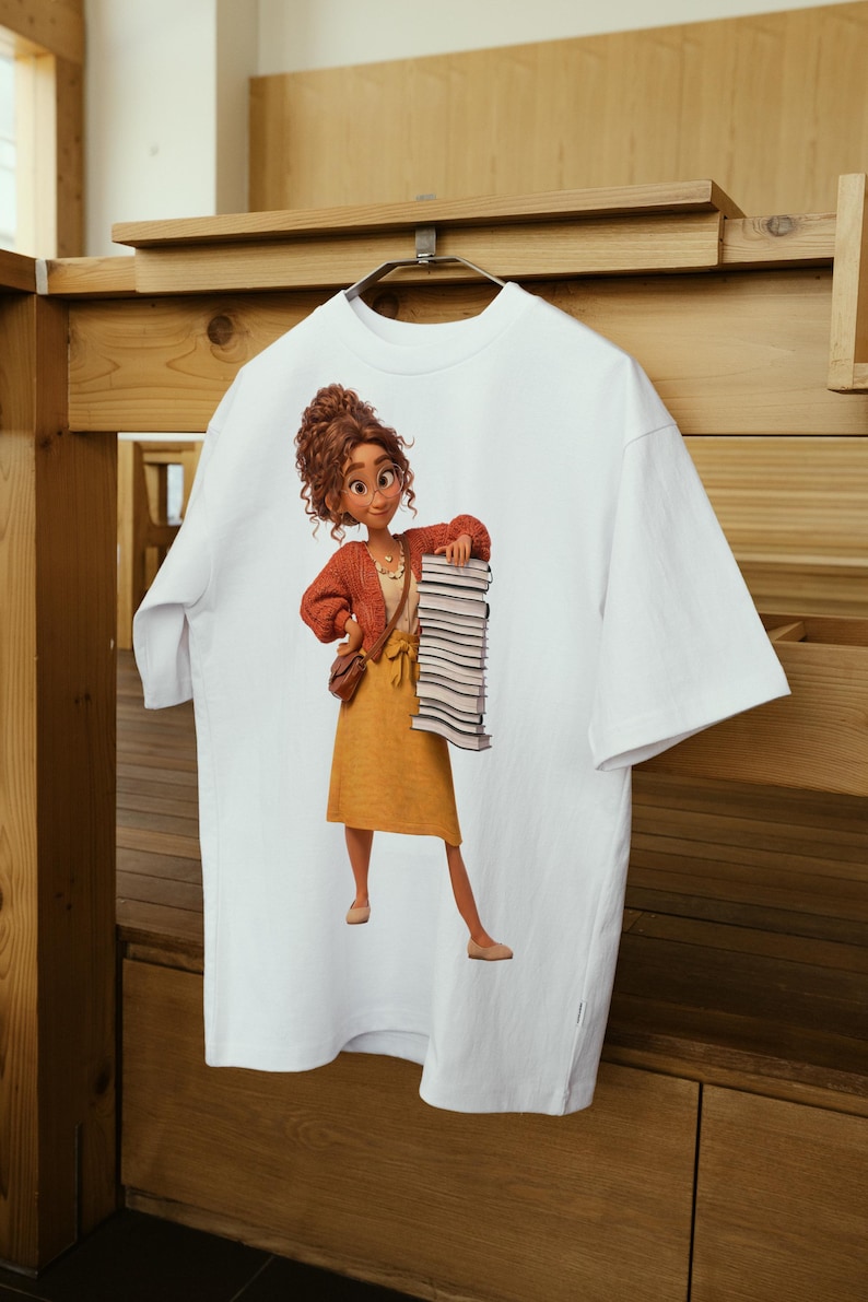 Peut inclure: Un t-shirt blanc &agrave; manches courtes avec une impression graphique repr&eacute;sentant une personne aux cheveux bruns boucl&eacute;s, portant un gilet rouille, une jupe jaune et des lunettes, tenant une haute pile de livres et un petit sac &agrave; bandouli&egrave;re marron.