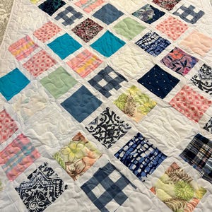 May include: A handmade quilt with a white background and a diamond pattern. Each diamond is a different color and pattern, including floral, plaid, and solid colors. The quilt is quilted with a wavy stitch.