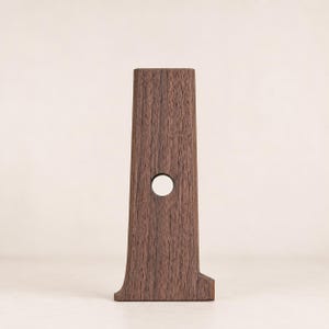 Könnte beinhalten: Ein dunkelbraunes Holzobjekt in Form der Zahl Eins, das ein vertikales, leicht verjüngtes Design mit einem kreisförmigen Ausschnitt in der Mitte aufweist. Die Holzmaserung ist auf der gesamten Oberfläche der aufrechten Struktur sichtbar.