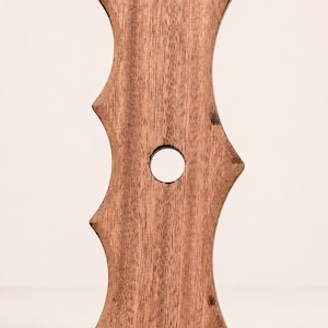 Könnte beinhalten: Ein vertikales, braunes Holzobjekt mit flacher Ober- und Unterseite, das eine symmetrische, wellenförmige Silhouette mit zwei spitzen Vorsprüngen auf der linken Seite und einem auf der rechten Seite aufweist. Ein kreisrundes Loch ist mittig.