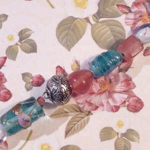 May include: A turquoise and pink beaded charm bracelet with a silver charm. The bracelet is on a floral patterned background.