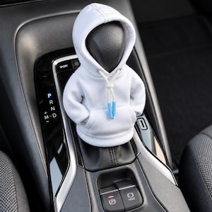 Könnte beinhalten: Ein weißer Miniatur-Kapuzenpullover als Überzug für einen Auto-Schaltknauf. Der Hoodie verfügt über eine Kapuze mit weißen Kordeln und hellblauen Kunststoffenden, eine Kängurutasche auf der Vorderseite und Ärmel, passend über dem Schalthebel.