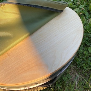 Könnte beinhalten: Ein rundes, helles Holzbrett, das teilweise in einer olivgrünen Tragetasche zu sehen ist. Die Tasche hat einen schwarzen Reißverschluss und ein grünes Innenfutter. Das Brett scheint aus mehreren Holzschichten zu bestehen.