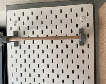 Könnte beinhalten: Eine weiße Lochwand mit gleichmäßig verteilten Löchern, einem Holzdübel und grauen Halterungen. Die Lochwand ist auf einem grauen Rahmen montiert, mit einer hellen Holzplatte auf der rechten Seite. Der Dübel ist etwa 30 cm lang.