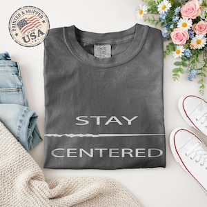 May include: A charcoal grey crewneck t-shirt featuring the text STAY CENTERED in white capital letters, separated by a thin horizontal line. The shirt has a Comfort Colors label at the inner neckline and is displayed with denim jeans and white sneakers.