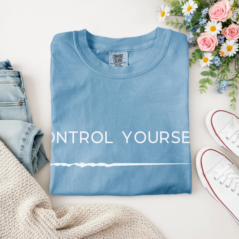 May include: A light blue Comfort Colors t-shirt with the words "CONTROL YOURSELF" printed in white. The shirt is folded and surrounded by white sneakers, blue jeans, and a beige sweater. Flowers are in the background.