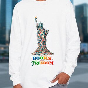 Op de afbeelding: Wit shirt met lange mouwen met een afbeelding van het Vrijheidsbeeld gemaakt van boeken. De woorden "BOOKS. FREEDOM" staan in kleurrijke letters onder de afbeelding. Het shirt is een comfortabele, casual stijl.
