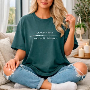 May include: Teal-colored t-shirt with the text "MASTER YOUR MIND" in white. The shirt is a relaxed fit, with short sleeves. The model is wearing blue jeans. The overall aesthetic is casual and comfortable.
