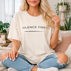 May include: Cream-colored t-shirt with the text "SILENCE FIRST" in black, above a horizontal line. The shirt is paired with light blue, distressed jeans. The overall aesthetic is casual and comfortable.