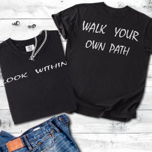 May include: Two black t-shirts with white text. One shirt says "LOOK WITHIN" and the other says "WALK YOUR OWN PATH." A silver chain necklace and blue jeans are also visible.