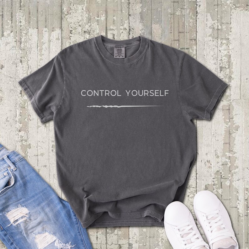May include: A charcoal gray short-sleeve t-shirt featuring the white text "CONTROL YOURSELF" printed on the chest above a thin, horizontal white line. The garment has a crew neckline and a relaxed fit with a visible brand label at the neck.
