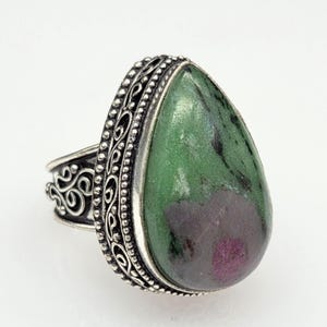 May include: A silver ring featuring a large, pear-shaped green and purple stone. The metal setting is decorated with intricate scrollwork and a beaded border surrounding the gemstone. The band displays detailed metalwork along the sides.