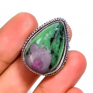 Puede incluir: Un anillo en forma de lágrima con una piedra preciosa grande. La piedra presenta colores verdes, negros y morados, engastada en una banda de metal plateado con un borde de cuentas. El anillo está sujeto por una mano.
