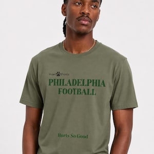 Op de afbeelding: Een olijfgroen t-shirt met korte mouwen en donkergroene tekst met de woorden Howl Prints, PHILADELPHIA FOOTBALL en Hurts So Good. Het shirt heeft een ronde hals en is gemaakt van een effen stof met een gezeefdrukt grafisch ontwerp.