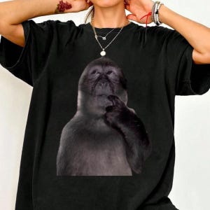 Puede incluir: Camiseta negra con un gráfico de un mono pensativo. El mono está representado en tonos grises y negros, con un dedo en la barbilla. La camiseta es de estilo casual y de gran tamaño.