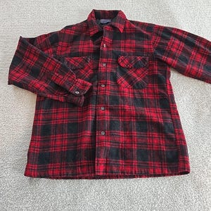 May include: A red and black plaid flannel shirt with a button-down front, two chest pockets, and long sleeves. The shirt is made of a soft, woven fabric and is laid flat on a light-colored surface.