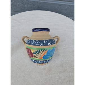 Op de afbeelding: Een kleine, handbeschilderde keramische vaas met twee handvatten. De vaas heeft een bruinachtige basis, een donkerblauwe rand en een decoratieve band met blauwe en witte krullen. De body is versierd met kleurrijke bloemmotieven in rood, blauw en groen.