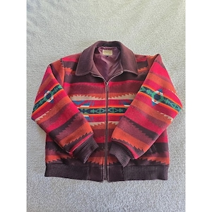 May include: A zip-up jacket with a Southwestern-inspired pattern. The jacket features a collar, ribbed cuffs, and a hem in a dark brown color. The body of the jacket is a mix of red, orange, and brown stripes with geometric designs in blue, green, and white.