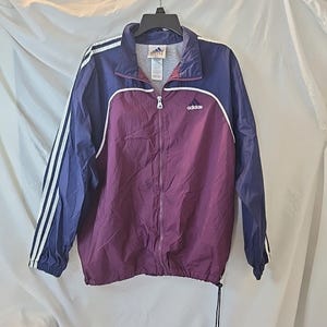 May include: A vintage Adidas track jacket in a color block design. The jacket features a deep purple body with navy blue sleeves and shoulders. White stripes accent the sleeves, and the Adidas logo is visible on the chest. A full zipper and drawstring hem complete the design.
