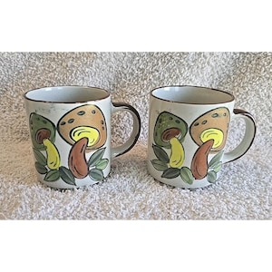 May include: Two ceramic mugs with a cream-colored base and brown trim. Each mug features a colorful mushroom design with green, yellow, and brown accents. The mugs have a curved handle and are suitable for hot beverages.