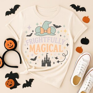 May include: Cream-colored t-shirt with a whimsical Halloween design. The shirt features a witch hat, bats, stars, and the words "Frightfully Magical." Decorative pumpkins, bats, and a pair of white sneakers complete the festive arrangement.