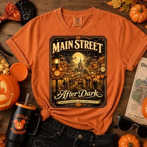 May include: An orange short-sleeve t-shirt featuring a graphic of a street scene at night with the text "MAIN STREET After Dark" and "WHERE HALLOWEEN MAGIC COMES TO LIFE". The design includes a full moon, a castle, and glowing street lamps.