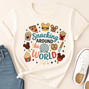 May include: A cream-colored short-sleeve t-shirt featuring a colorful graphic print of various snacks and the text "Snacking AROUND the WORLD" with a central geodesic dome icon, surrounded by small star accents and food illustrations.
