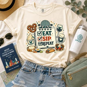 May include: A cream-coloured short-sleeve t-shirt featuring a graphic of a checklist with the text EAT, SIP, REPEAT. The design includes illustrations of a pretzel, soft serve, dumplings, sushi, a taco, waffles, and drinks, all surrounding the text.