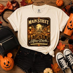 May include: A cream-colored t-shirt featuring a graphic print of a nighttime street scene with a castle, a full moon, and the text MAIN STREET After Dark WHERE HALLOWEEN MAGIC COMES TO LIFE, surrounded by a black backpack, sneakers, and pumpkins.