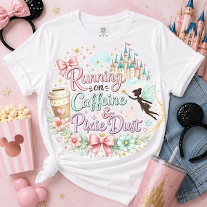 May include: A white short-sleeve t-shirt featuring a graphic print with a castle, a coffee cup, a fairy, and bows. The text reads Running on Caffeine & Pixie Dust. The design includes floral accents, stars, and sparkles in pastel colors.