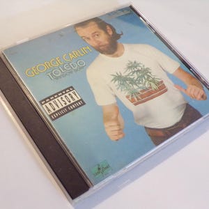 May include: A compact disc case with a blue background featuring a man wearing a white t-shirt with a marijuana plant graphic. The text "George Carlin Toledo" and "Parental Advisory Explicit Content" is printed on the case.