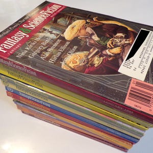 May include: A pile of vintage "Fantasy & Science Fiction" magazines. The top magazine has a fantasy-themed cover illustration, with the title in red and green. The magazines are stacked, with visible dates and titles.