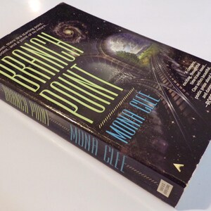 May include: A hardcover book titled "Branch Point" by Mona Clee. The cover features a dark, starry background with a railway track leading towards a bright, surreal landscape. The title is in large, neon green letters.