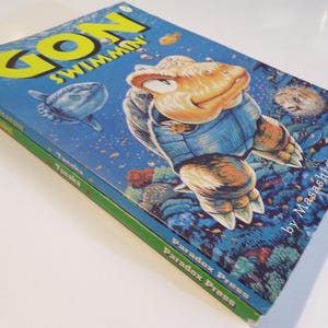 Könnte beinhalten: Zwei gestapelte Comicbücher mit dem Titel "GON SWIMMIN" mit blauem Cover und einer Zeichentrickschildkröte. Das untere Buch hat einen grünen Buchrücken und den Text "Paradox Press". Der Name des Autors, Masashi Tanaka, ist ebenfalls sichtbar.