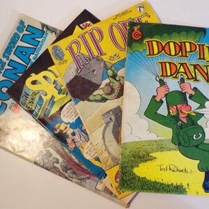 Puede incluir: Tres cómics vintage, uno con una portada azul con el texto "The Savage Sword of Conan", otro con una portada amarilla con el texto "Rip Off" y un tercero con una portada azul y verde con el texto "Dopin' Dan".