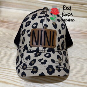 May include: A trucker hat with a leopard print pattern in tan and black. The hat features a black mesh back and a brown leather patch with the name "NINI" in black letters. The Red Rose Keepsakes logo is visible on the right side.