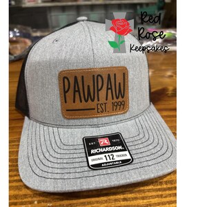 May include: A gray and black trucker hat with a brown leather patch that reads "PAWPAW EST. 1999". The hat has a curved brim and an adjustable strap. The hat also has a tag that reads "RICHARDSON ORIGINAL 112 TRUCKER ADJUSTABLE".