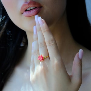 May include: A gold ring with a pink flower design. The ring's petals are a vivid pink, and the band is gold. The ring is worn on a finger, highlighting its delicate floral details. A close-up shot.