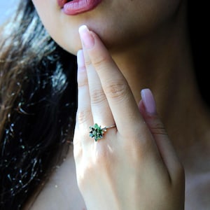 May include: A silver ring with a floral design, set with a cluster of green gemstones. The gemstones are a rich emerald green, and the band is polished silver. The ring is displayed on a finger.