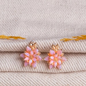 May include: A pair of gold-coloured stud earrings in a flower shape. Each earring has pink opal-like stones arranged around a central stone. The earrings have a detailed leaf design and small clear stones.